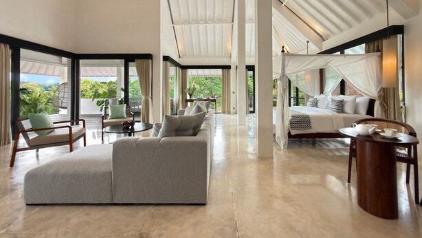 Amplia sala de estar con un sofá seccional gris grande, una mesa de centro redonda de madera y una cama con dosel. La habitación cuenta con ventanas amplias que ofrecen vistas a la vegetación exterior.