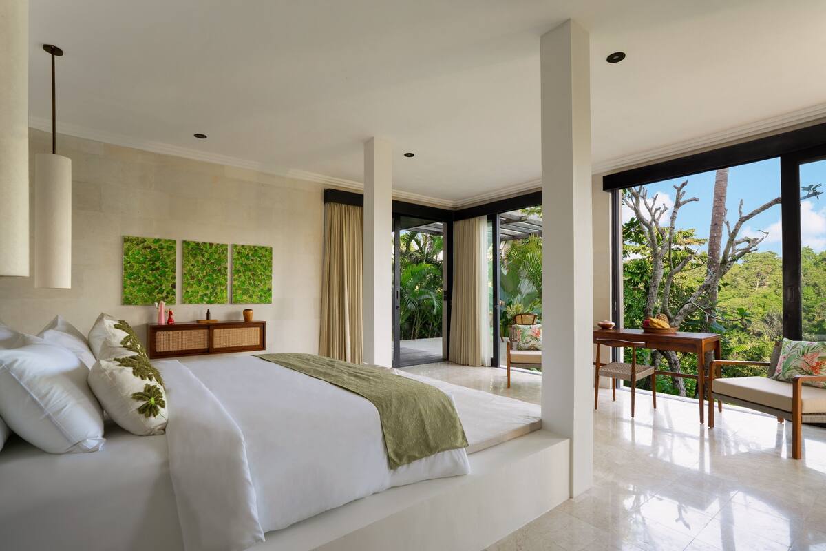 A modern bedroom with a large bed, a dining area with a table and chairs, and a view of greenery outside.