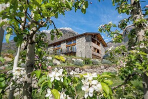 Une maison en pierre dotée d’un balcon en bois, entourée de montagnes et de plantes en fleurs.