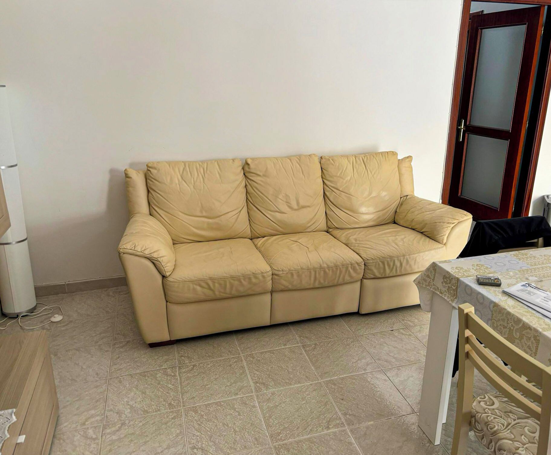 A beige leather sofa in a room with tiled flooring, a wooden door, and a small table with a patterned tablecloth.