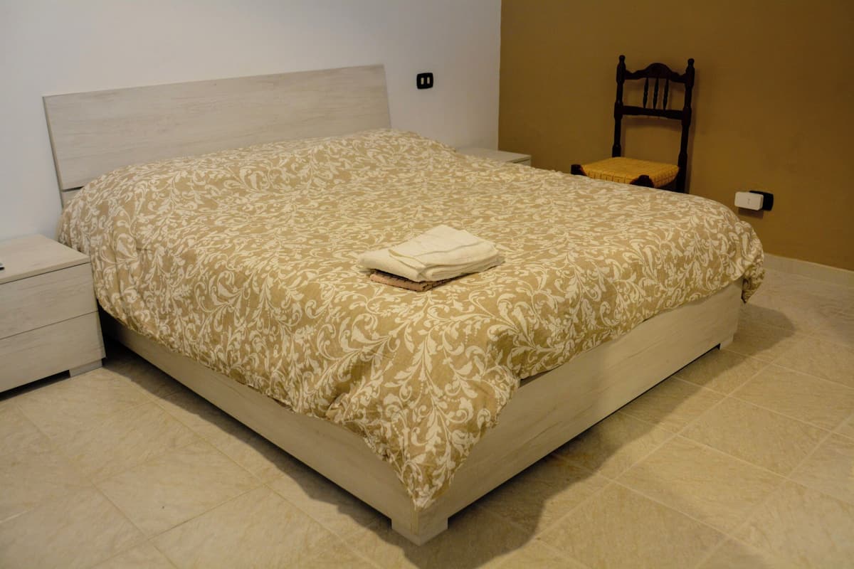 A neatly made bed with a patterned bedspread, a wooden nightstand, a chair, and a wall socket.