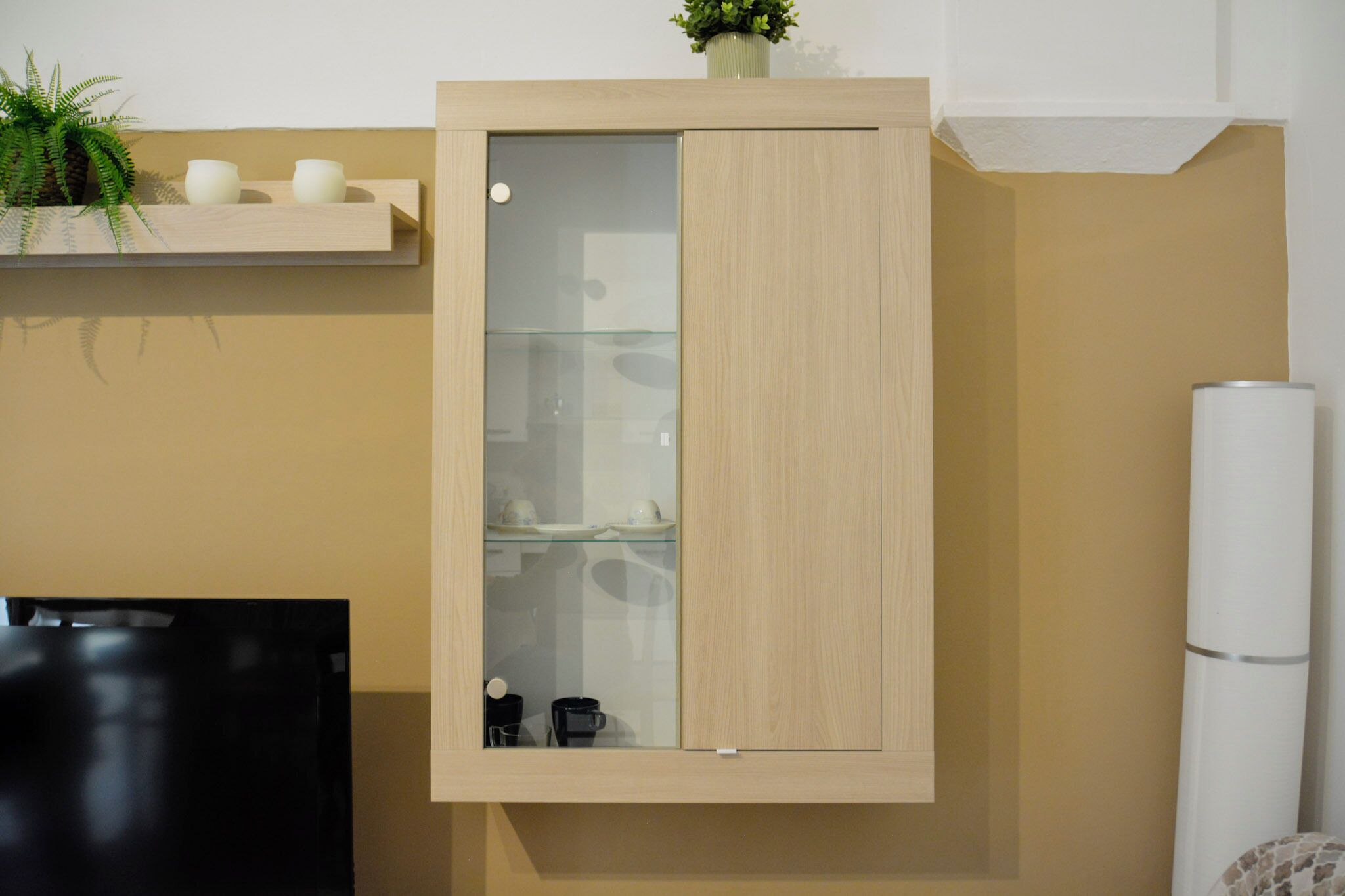A wooden cabinet with a glass door, a shelf with decorative items, and a white cylindrical object to the right.