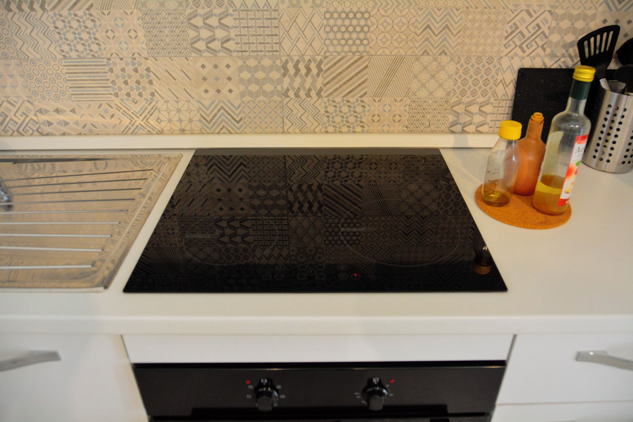 Una cocina con un salpicadero decorado, una cocina de inducción negra y diversos utensilios de cocina y botellas sobre la encimera.