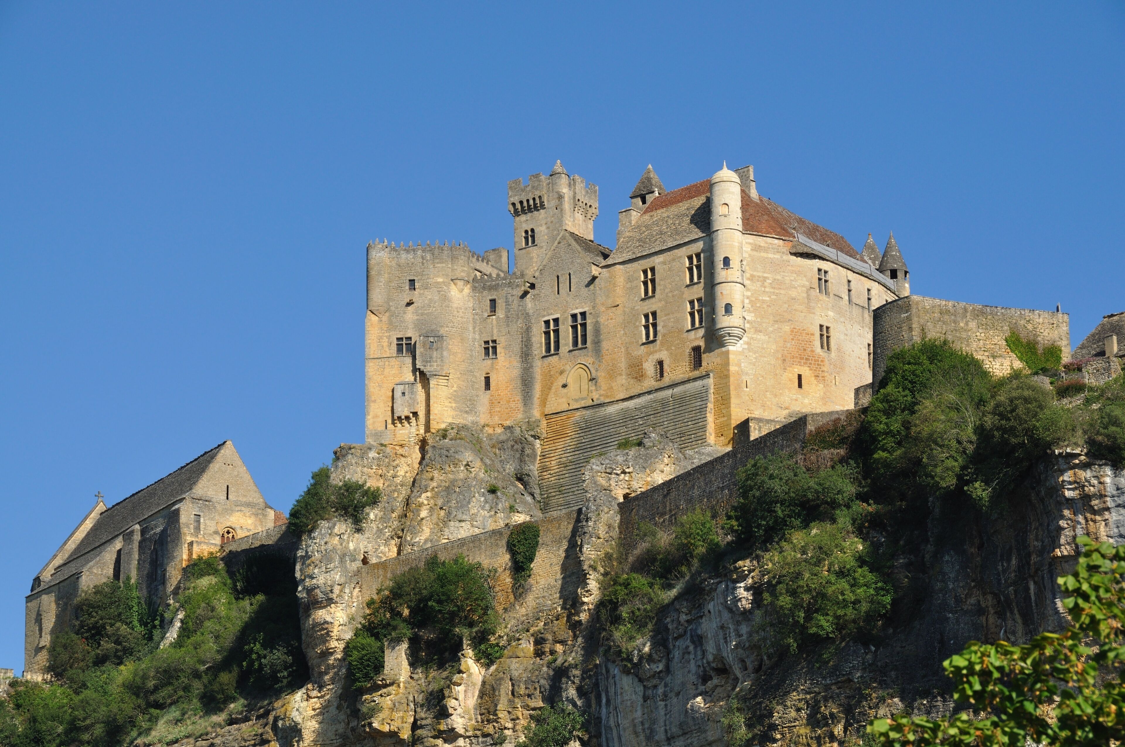 Eine historische Burg mit einem markanten Turm, gelegen auf einem felsigen Hügel inmitten von Grünflächen.