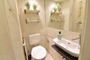 Ein Badezimmer mit Toilette, Waschbecken und Dusche. Es gibt Regale mit Pflanzen und einen Spiegel über dem Waschbecken.