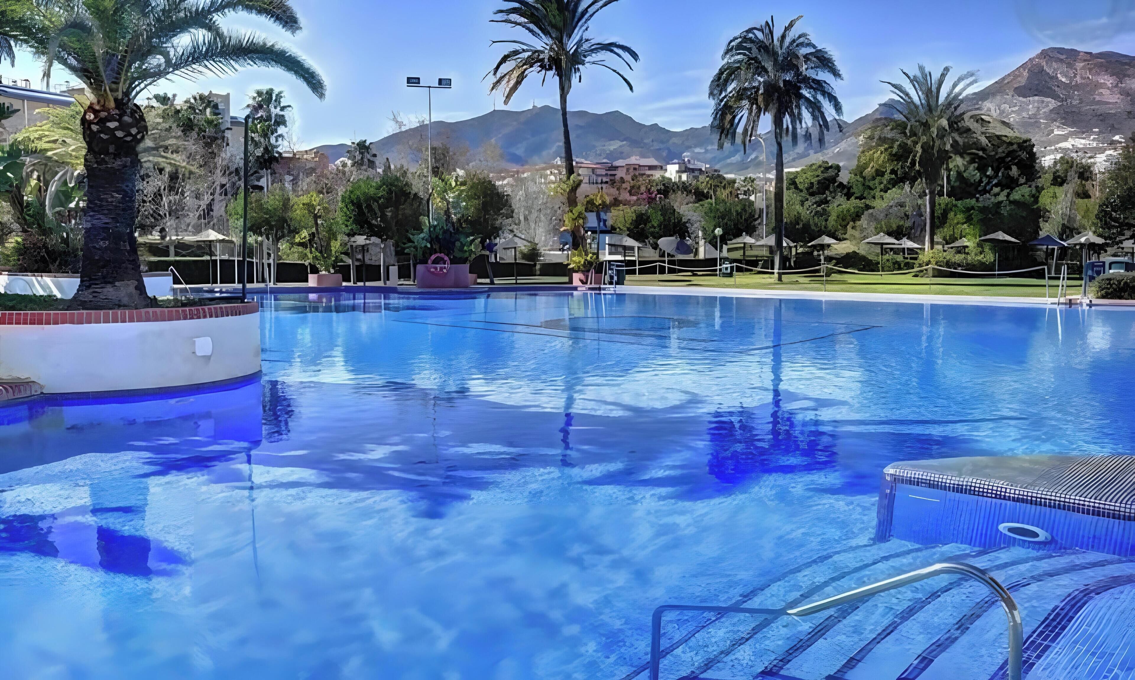 Una piscina con agua cristalina, rodeada de palmeras y montañas al fondo.