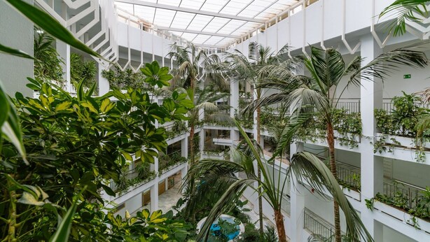 Ein mehrgeschossiges Atrium mit Glasdecke, umgeben von üppiger Vegetation und weißen Strukturelementen.