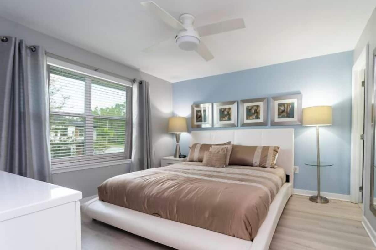 A bedroom with a bed, a ceiling fan, a bedside table with a lamp, and a window with curtains.