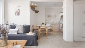 A modern living room with a sofa, coffee table, and a dining area with chairs. There is a kitchen in the background with cabinets and a sink. - Azure 2, Morfes Luxury Apartments (Tinos)