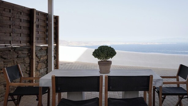A table with four chairs, a potted plant, and a view of a beach and mountains. - Azure 2, Morfes Luxury Apartments (Tinos)