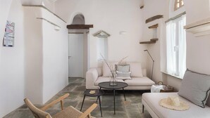 A cozy living room with a sofa, a coffee table, and a chair. There is a window with curtains, a wall-mounted shelf, and a decorative mirror. - Green 1, Morfes Luxury Apartments (Tinos)