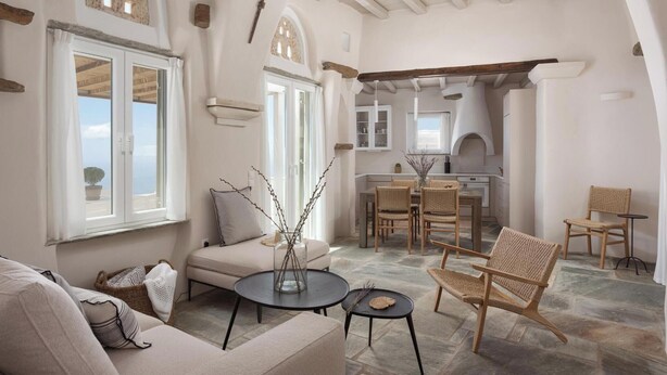 A spacious living area with a sofa, round coffee table, and wicker chairs. A dining table with chairs is in the background. The room features exposed wooden beams, white walls, and large windows. - Green 1, Morfes Luxury Apartments (Tinos)