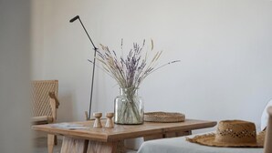 A wooden table with a glass vase containing dried plants, a woven chair, and a straw hat on a bed. - Green 3, Morfes Luxury Apartments (Tinos)