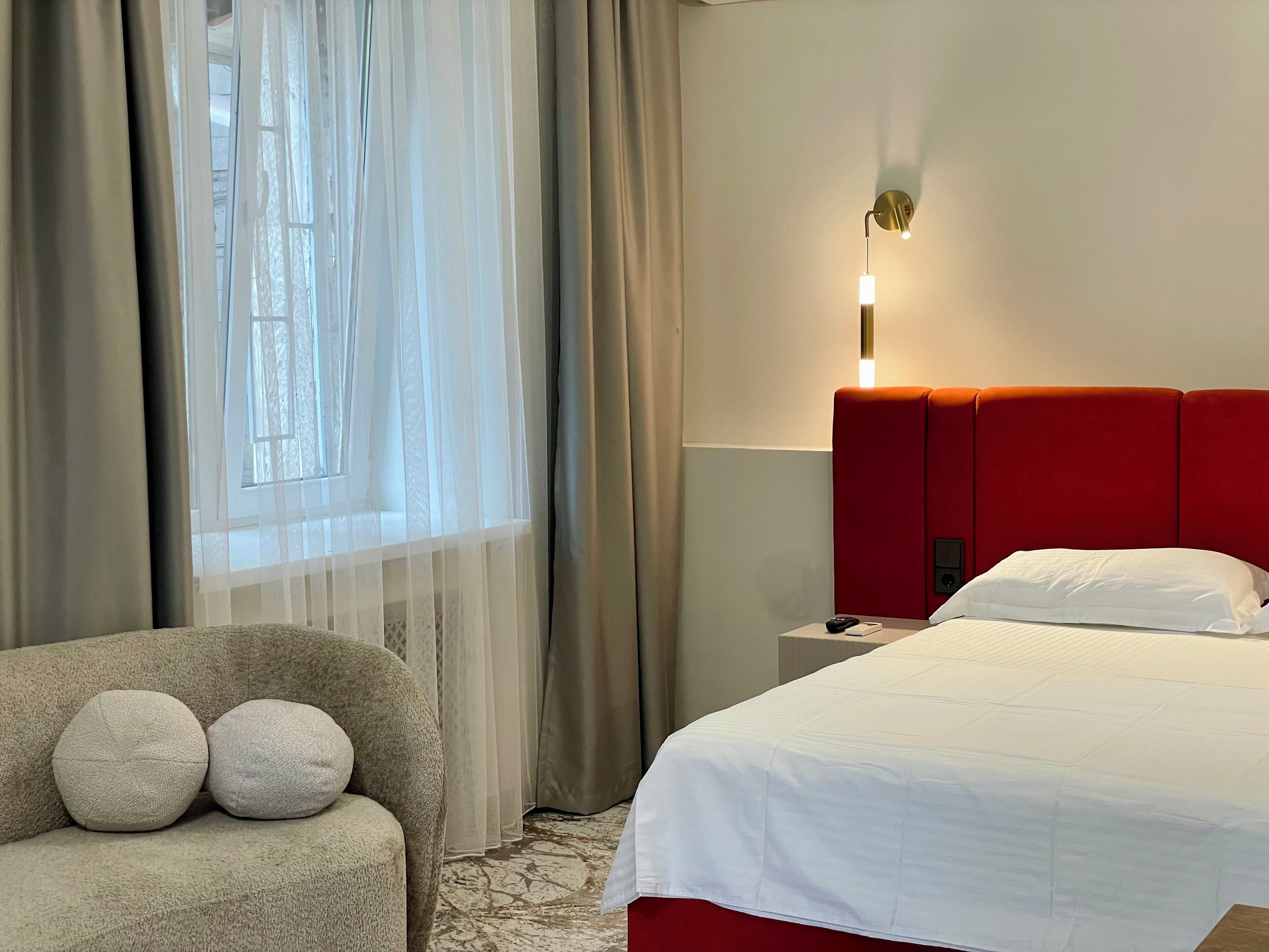 A hotel room with a bed, a nightstand, a lamp, a chair with cushions, and sheer curtains.