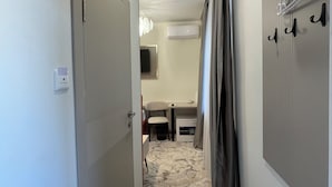 A narrow hallway with a door on the left, a bed with a headboard on the right, and a small table with a chair in the middle. - Naya Hotel (Almaty)