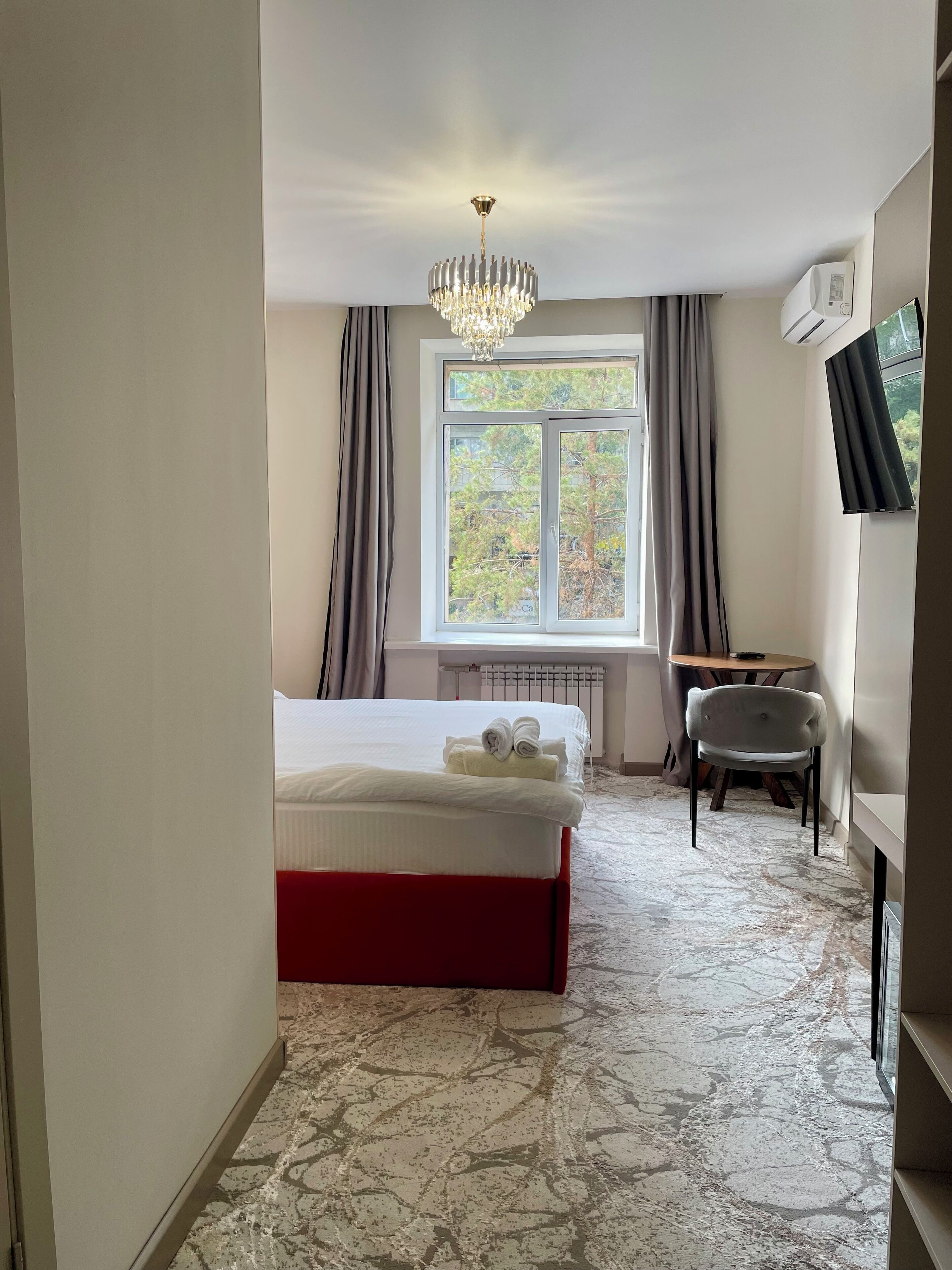 Ett hotellrum med en säng, ett skrivbord, en stol, ett fönster med gardiner och en kristallkrona.