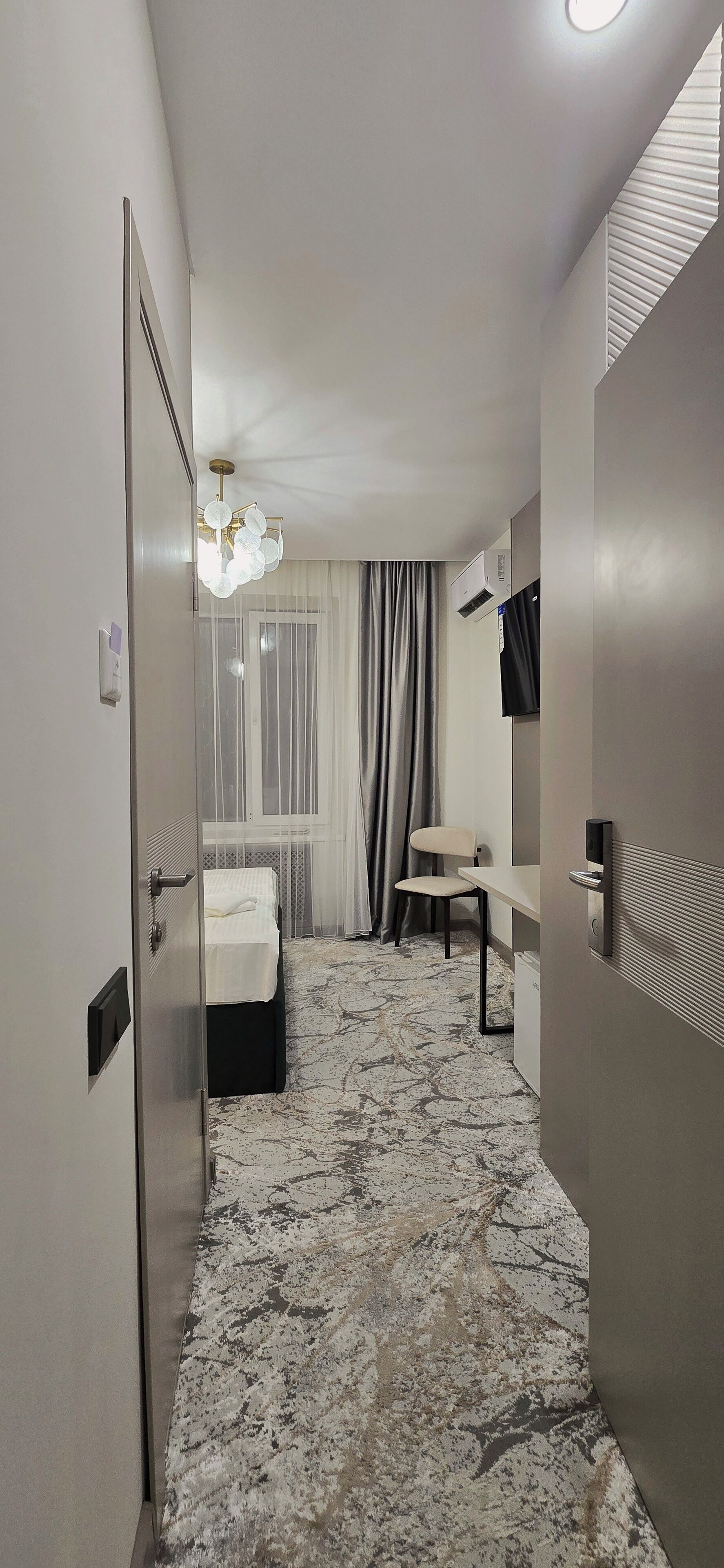 Una habitación de hotel con cama, una silla, un escritorio, televisor y ventana con cortinas.