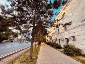 A paved pathway alongside a building with a stone facade, a road with vehicles, and trees. - Naya Hotel (Almaty)