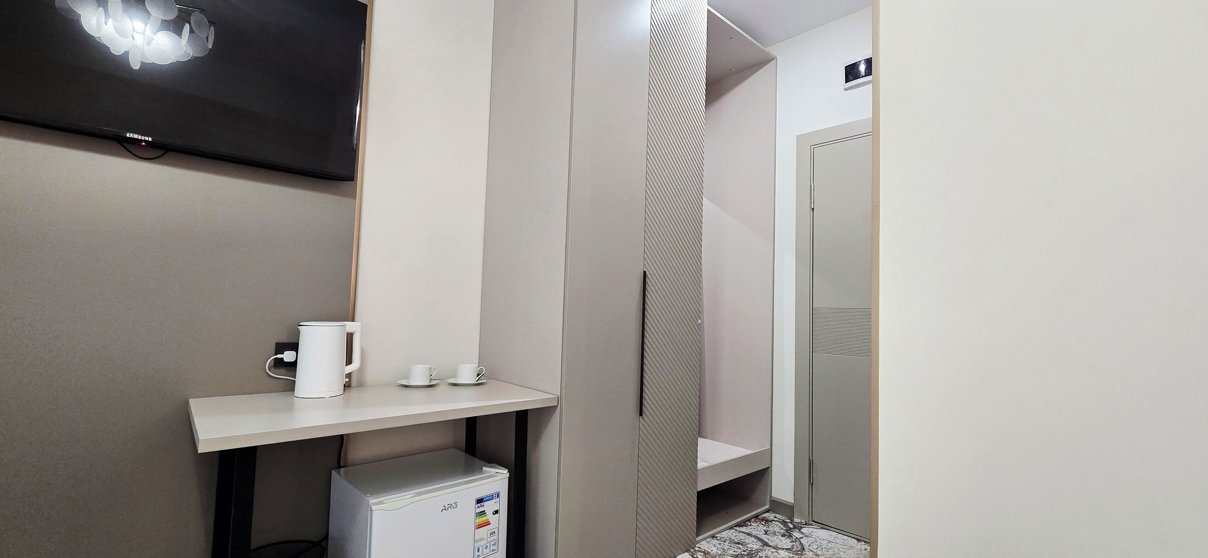 Una habitación de hotel moderna con una televisión de pantalla plana montada en la pared, una mesita con cafetera y tazas, un refrigerador pequeño y un armario.