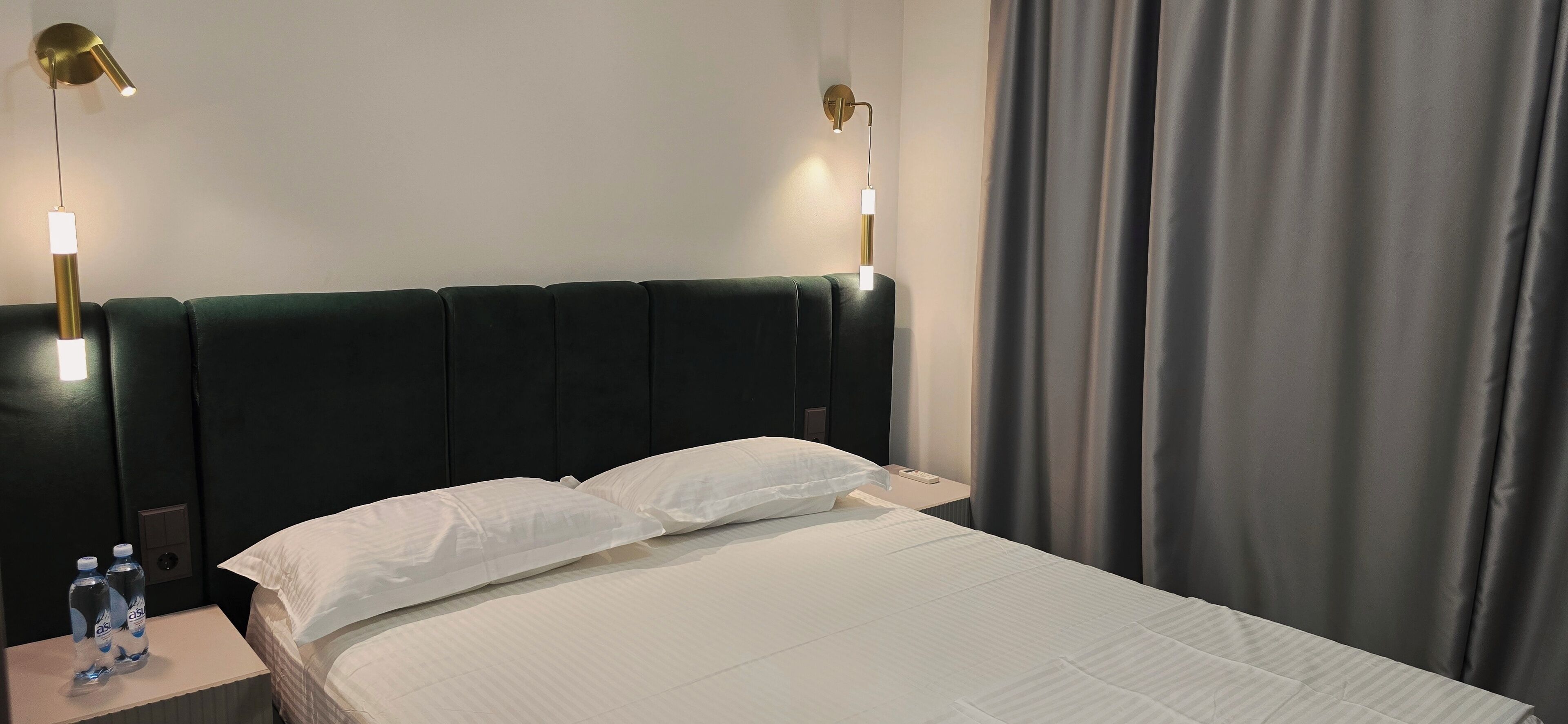 A hotel room with a bed, two bedside tables, a nightstand, a wall-mounted lamp, and a curtain.