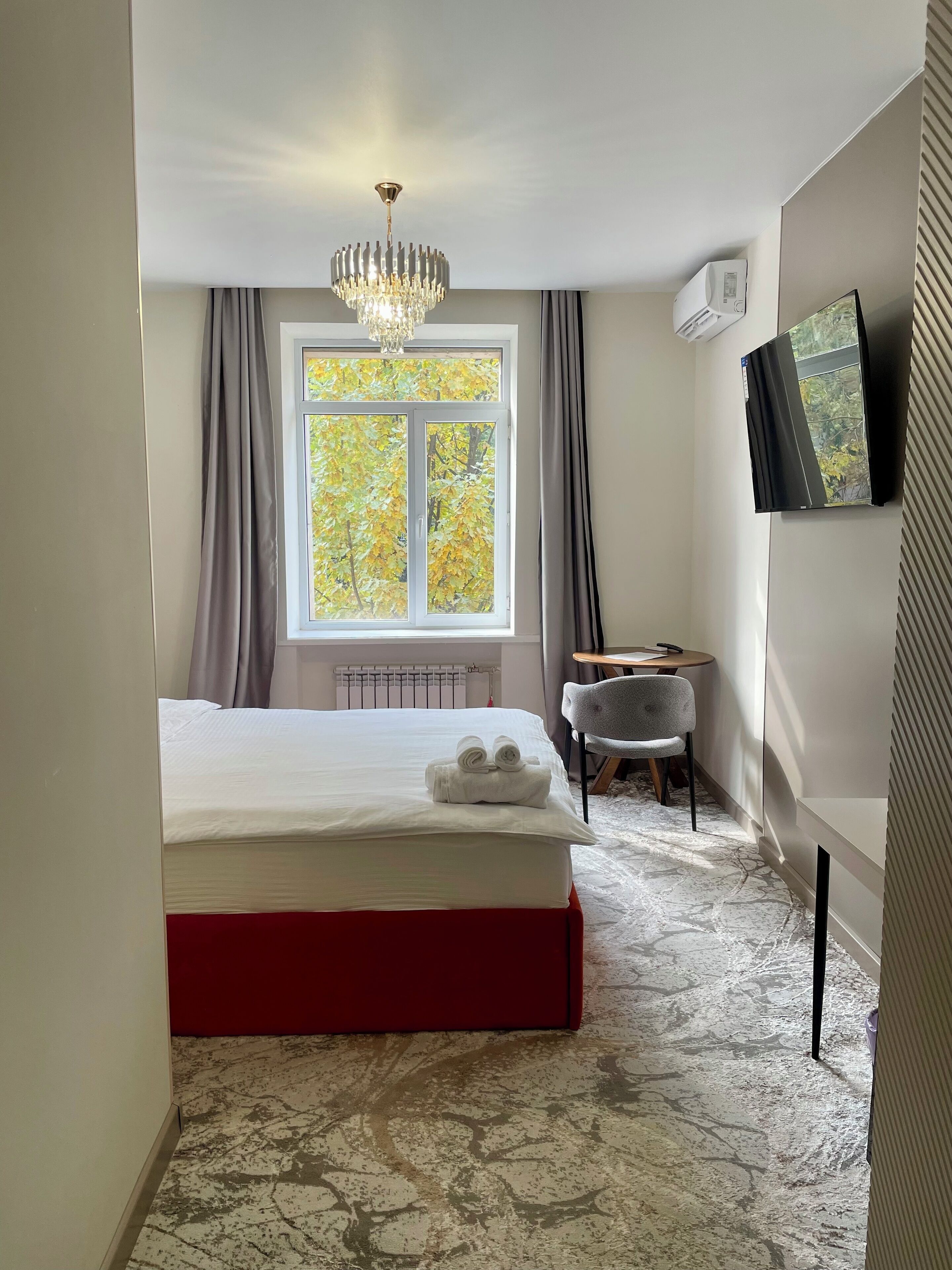 Habitación de hotel con cama, escritorio, silla, ventana con cortinas, candelabro y televisor de pared.