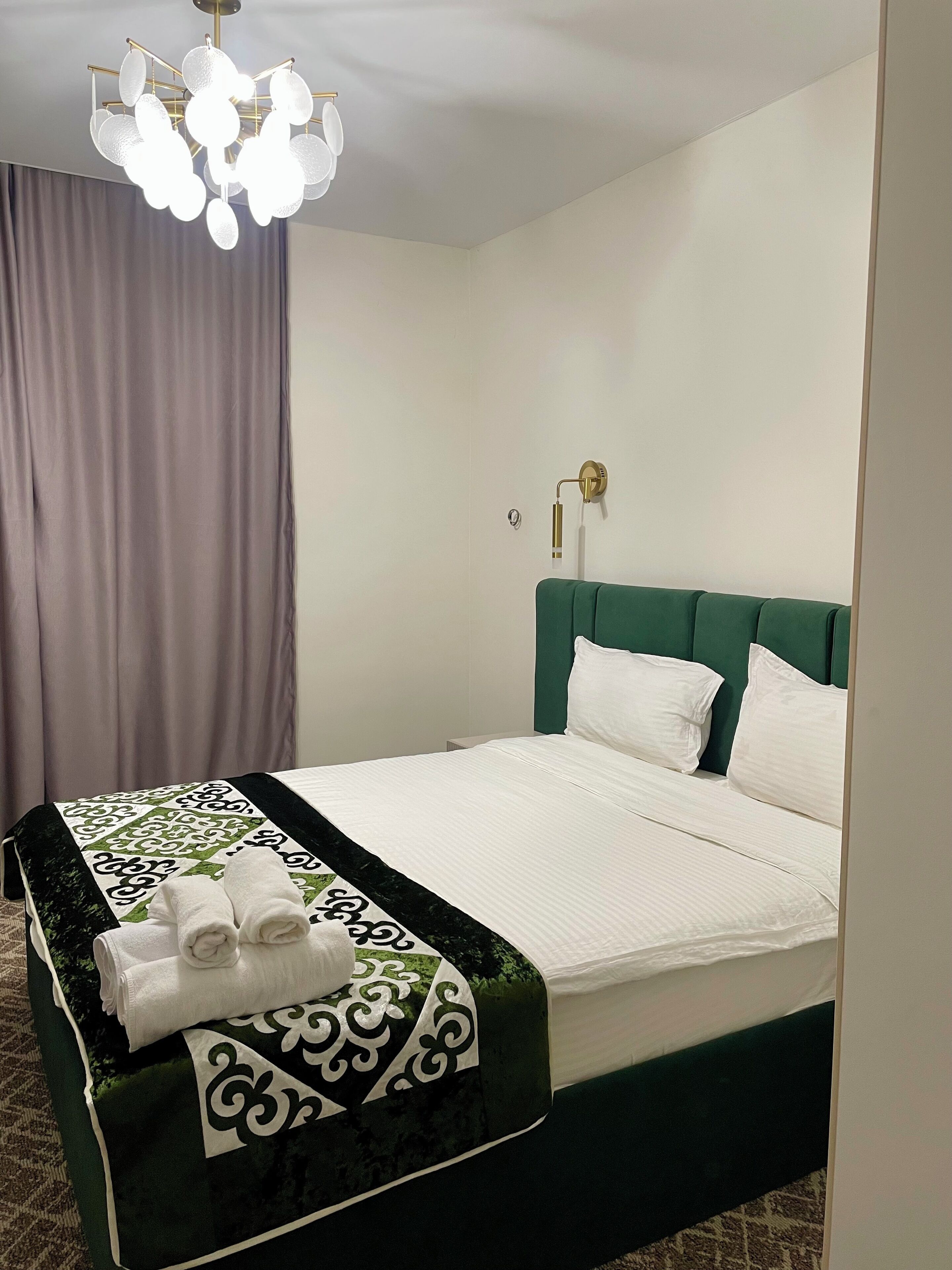 A neatly made bed with white linens and a green headboard, a decorative throw blanket, and a set of towels on a bedside table.