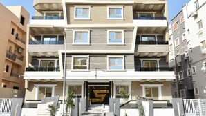 A multi-story hotel with a modern design, featuring balconies on each floor, a central entrance with a staircase, and a sign indicating the name "Stay Inn". - Stay Inn51 Al Sheikh Zayed (Giza Governorate)