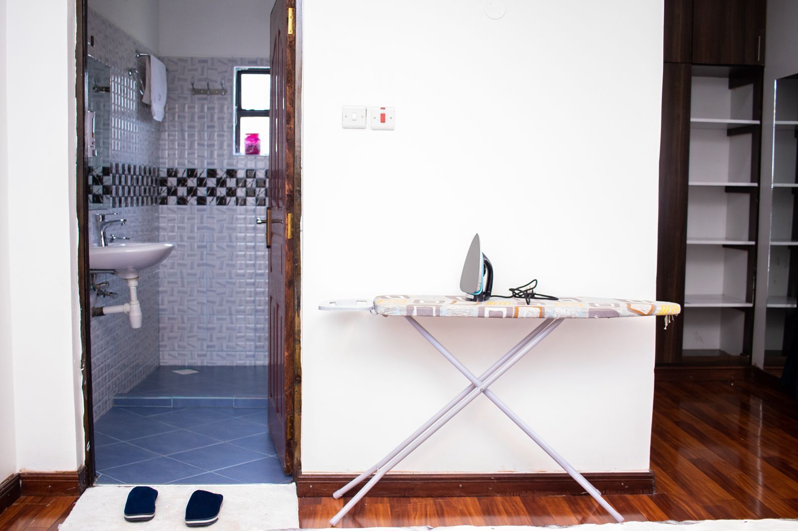 Un baño con lavamanos y ducha, una plancha con tabla para planchar pequeña, y un par de zapatillas en el suelo.