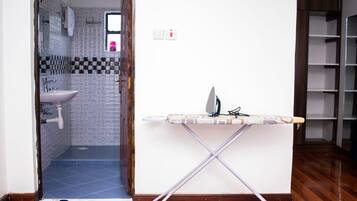 Un baño con lavamanos y ducha, una plancha con tabla para planchar pequeña, y un par de zapatillas en el suelo.