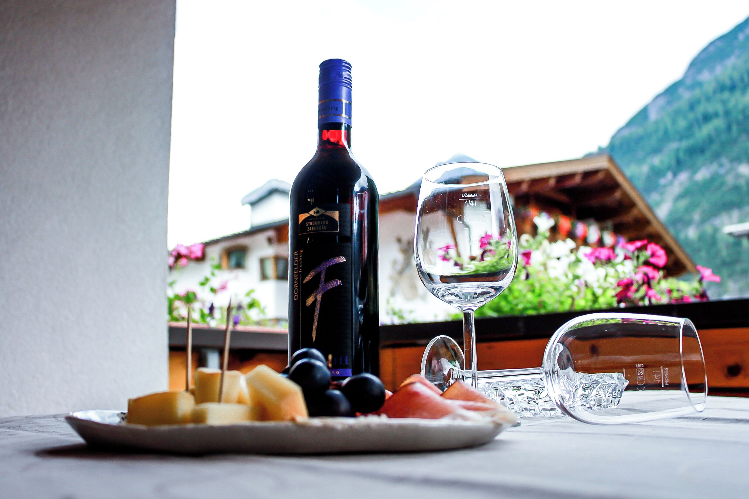 A wine bottle, a wine glass, and a plate with cheese and fruit on a table with a mountain view.