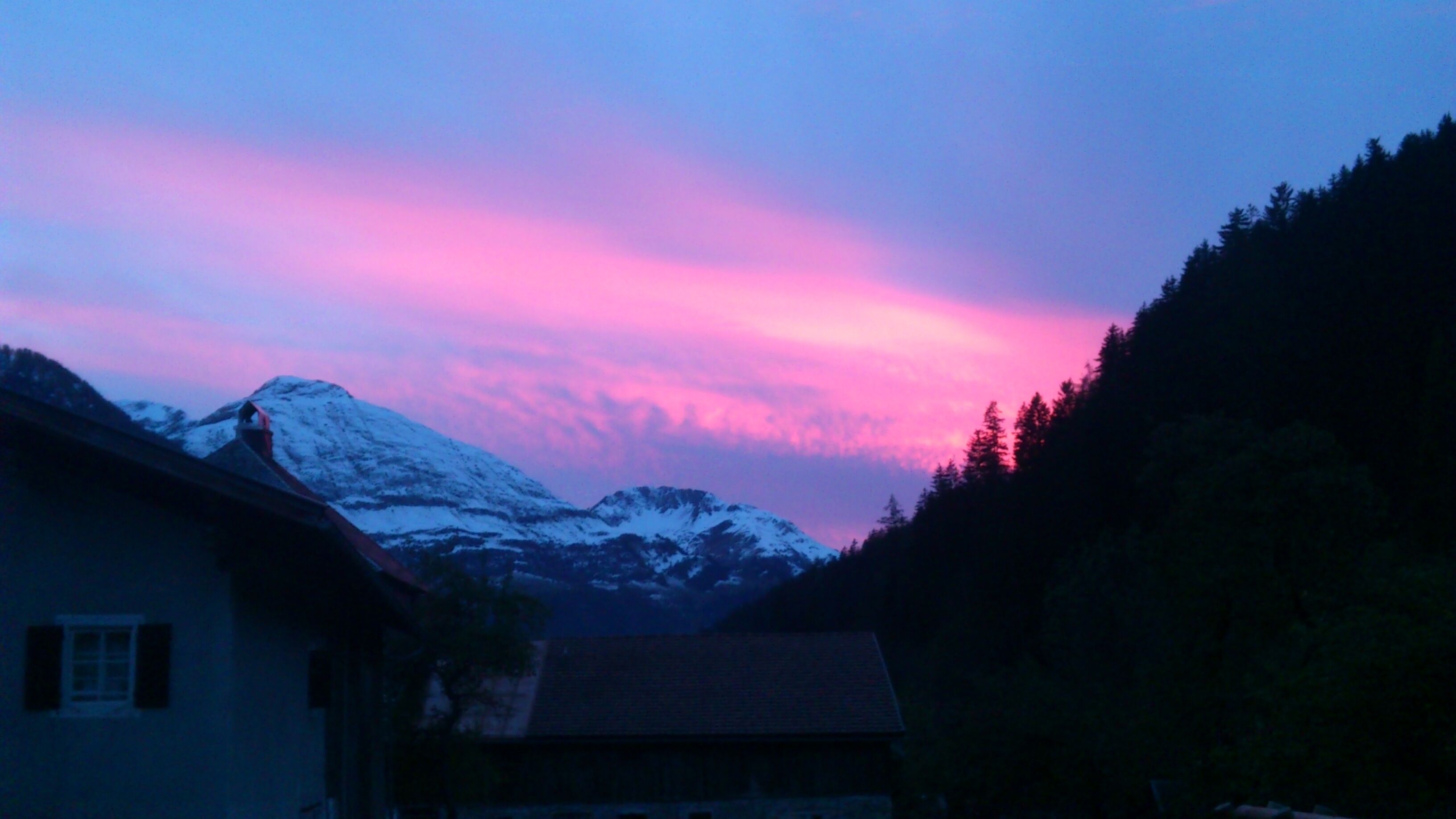 Une montagne dont le sommet est enneigé, une maison avec un toit en pente et une colline boisée.