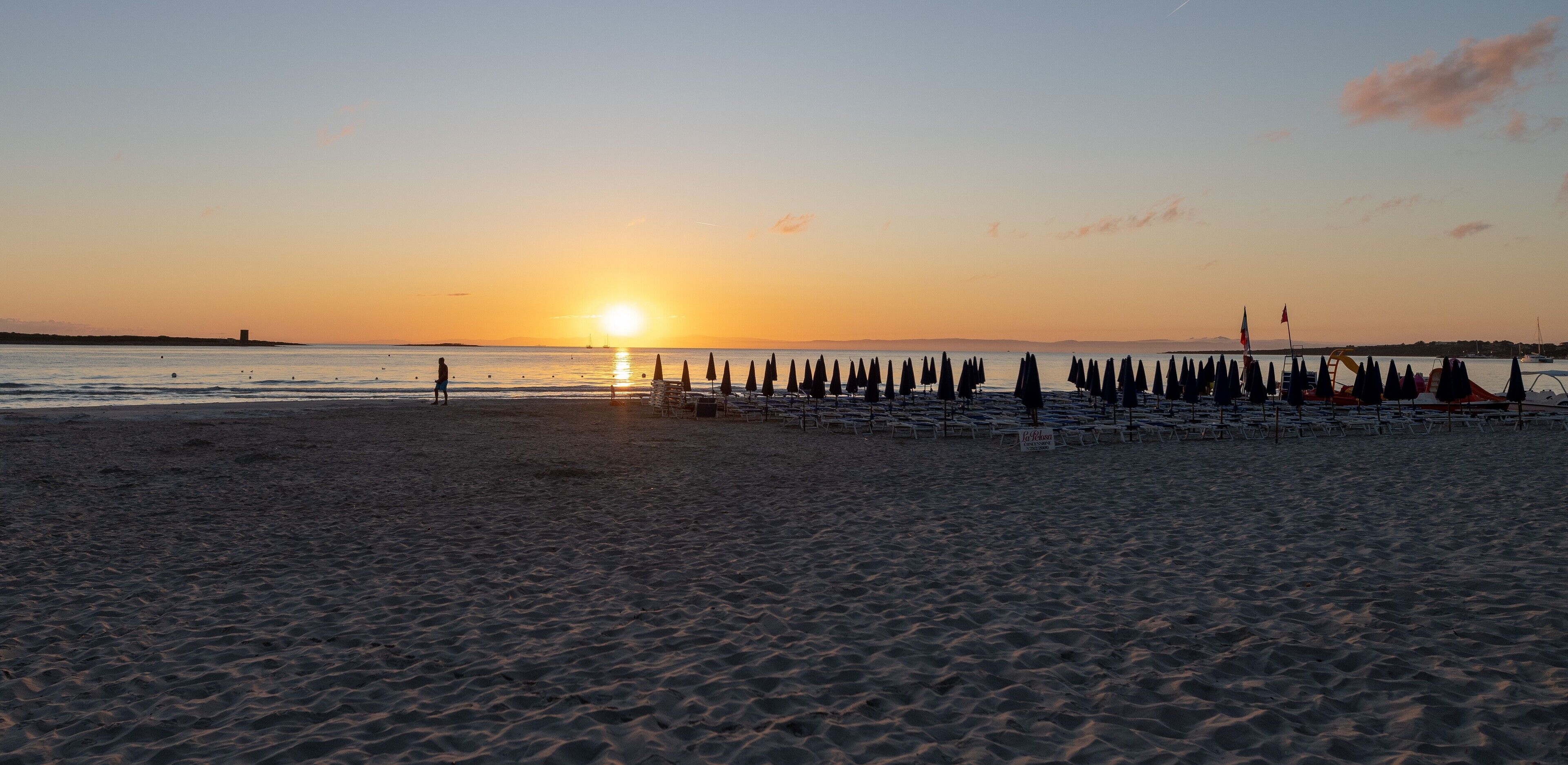 Una spiaggia con file di sedie a sdraio e ombrelloni, una persona che cammina e un tramonto sullo sfondo.