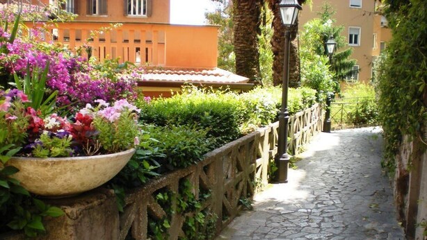 A cobblestone pathway flanked by a wooden fence and lush greenery, leading to a building with a terracotta roof. - House of alleys (Roma)