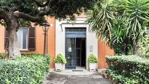 A building entrance with a paved path leading to a door, surrounded by greenery and a tree. - House of alleys (Roma)