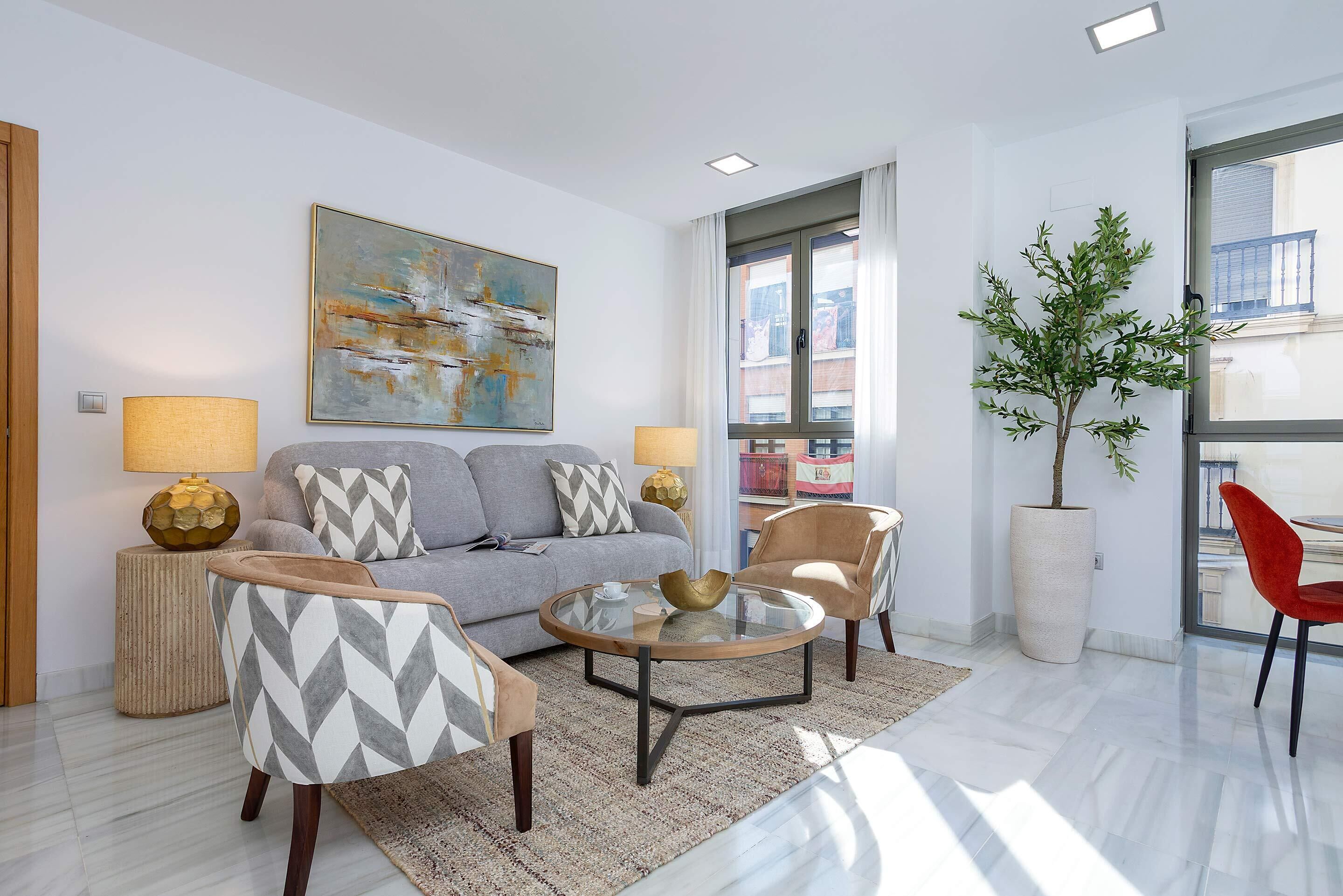 A modern living room with a grey sofa, patterned armchairs, a glass coffee table, and a large abstract painting.