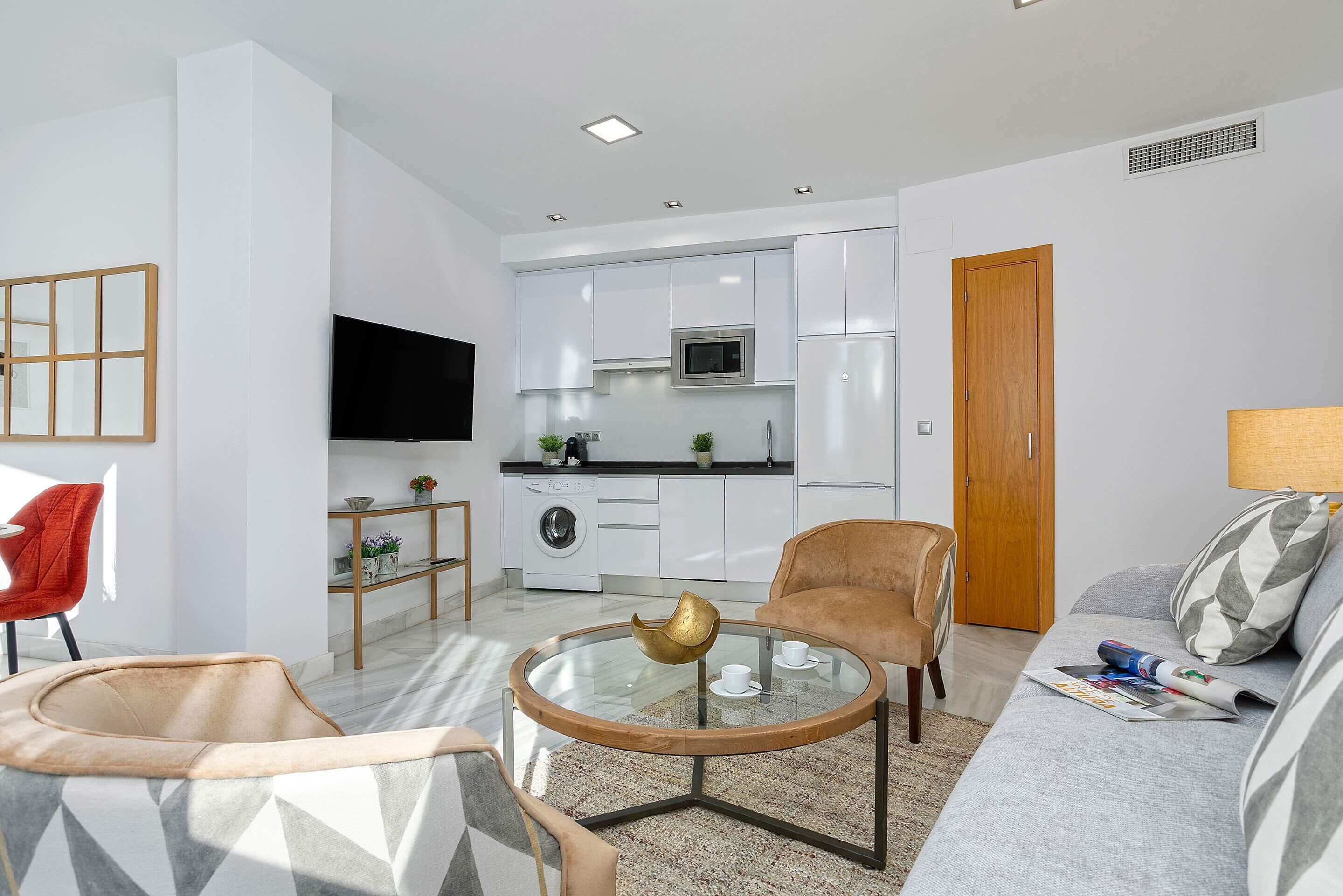 A modern living room with a glass coffee table, a sofa, a red chair, and a kitchen in the background.