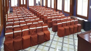 Une salle de conférence avec des rangées de chaises recouvertes d’un tissu marron, de grandes fenêtres en arc de cercle et des luminaires au plafond.
