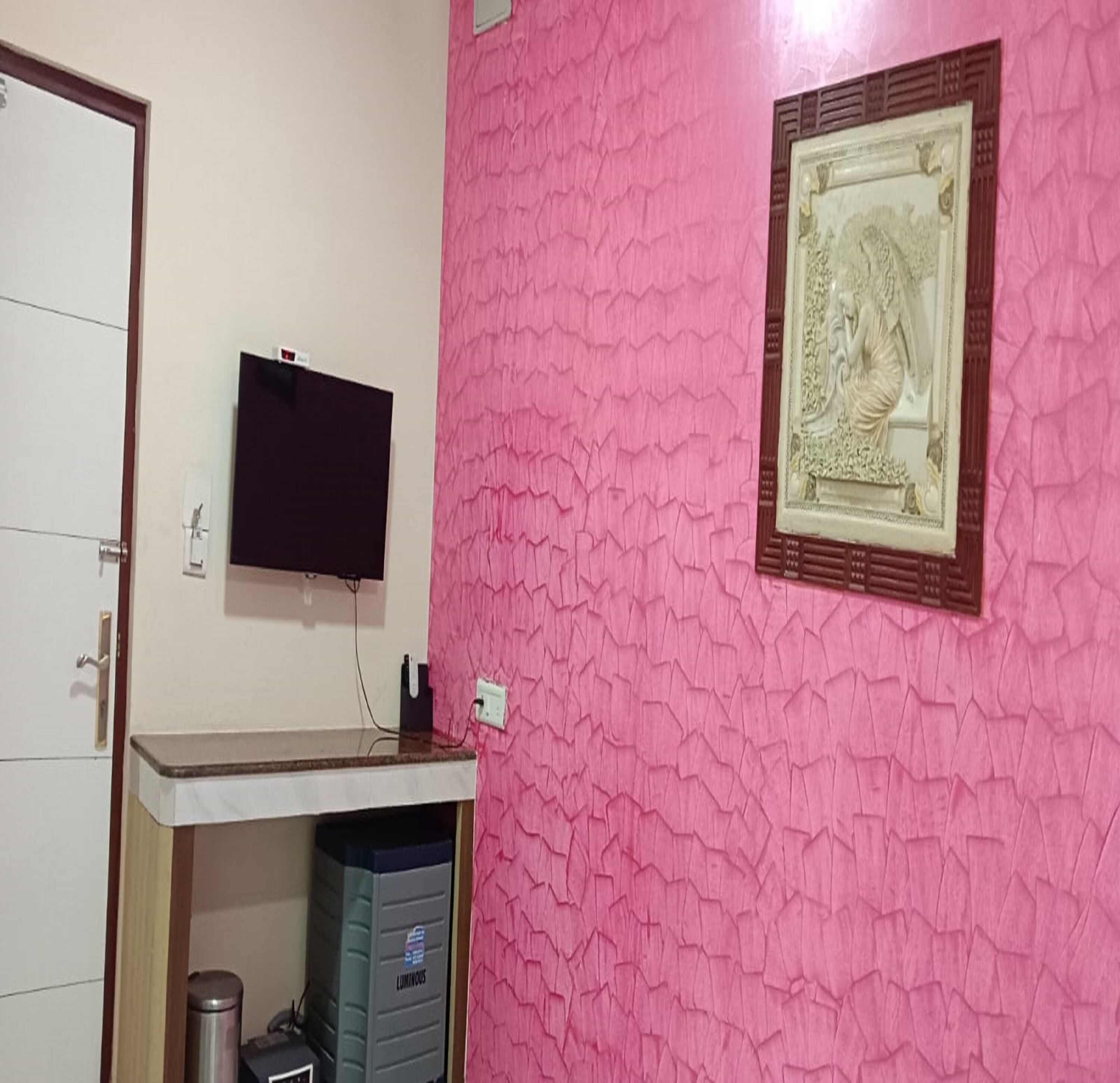 Una habitación con pared rosa texturizada, televisor montado, cuadro enmarcado, mueble blanco y un cubo de basura.