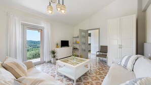 Ein geräumiges Wohnzimmer mit einer Couch, einem Couchtisch und einem Fernseher. Ein Balkon bietet Blick auf Grünflächen.