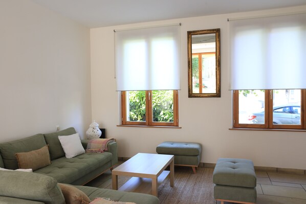 Una sala de estar con un sofá verde, una mesa blanca y un reposapiés verde. Hay dos ventanas con persianas, un espejo y una lámpara sobre una mesita auxiliar.
