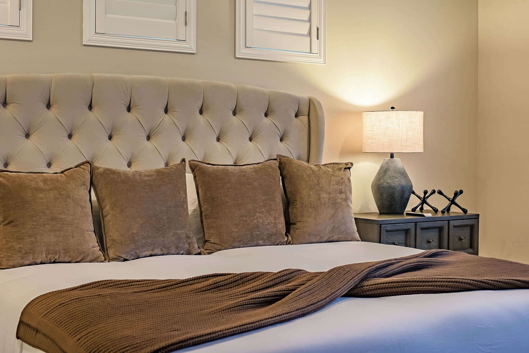 Un dormitorio con cabecera capitonada, cama con ropa de cama marrón, mesita de noche con lámpara y estante fijado a la pared.