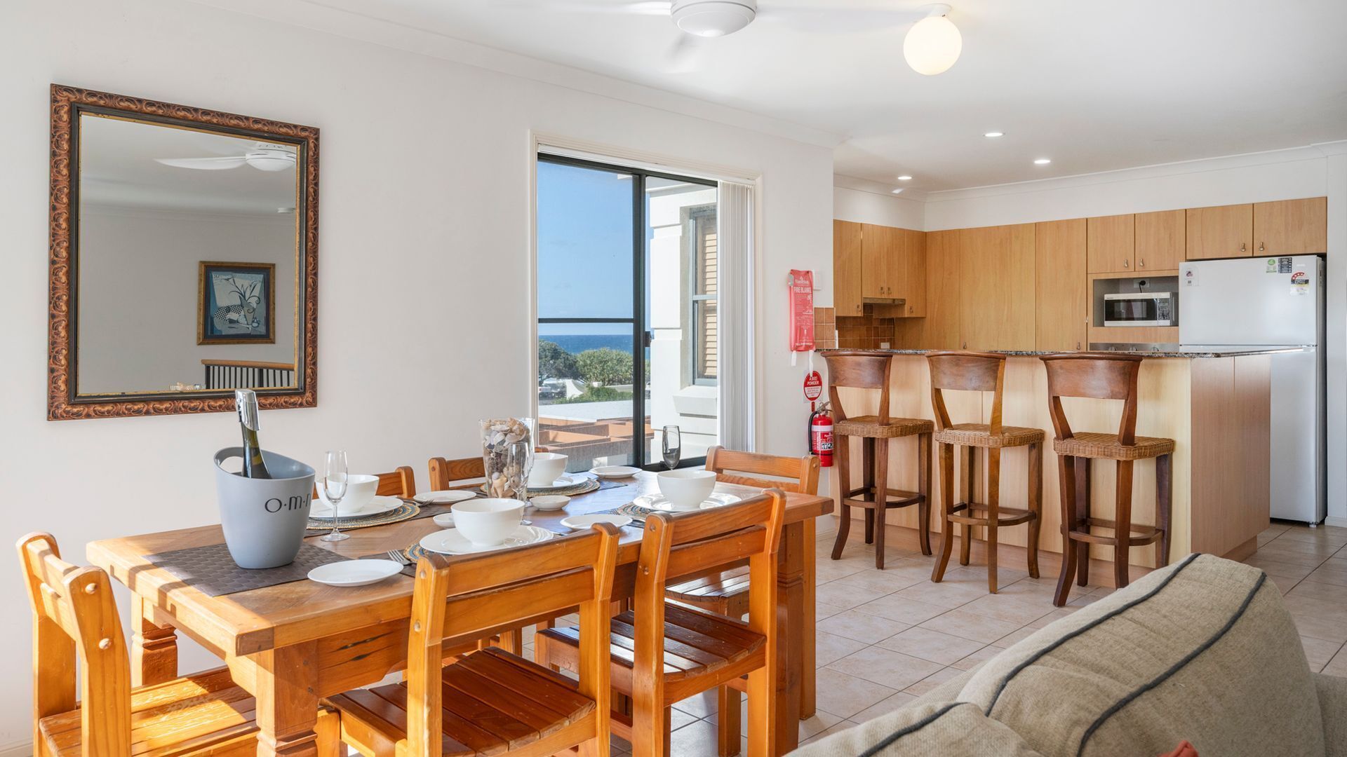 Un coin repas avec une table dressée pour quatre personnes, une cuisine dotée de meubles en bois et une vue sur la mer par une grande fenêtre.