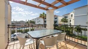 A balcony with a glass table and chairs, overlooking a residential area with palm trees and a swimming pool. - Breakers Block 3 Unit 2 - Lifestyle Yamba (YAMBA)