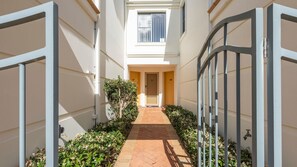 A narrow pathway leading to a building entrance with a metal gate, surrounded by greenery and a tiled floor. - Breakers Block 3 Unit 2 - Lifestyle Yamba (YAMBA)