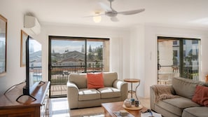 A spacious living room with a large window, a ceiling fan, and a view of a residential area. - Breakers Block 3 Unit 2 - Lifestyle Yamba (YAMBA)