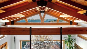 Ein Zimmer mit schrägverdeckter Holzbalkendecke, großen Fenstern und Blick auf eine Stadtlandschaft.