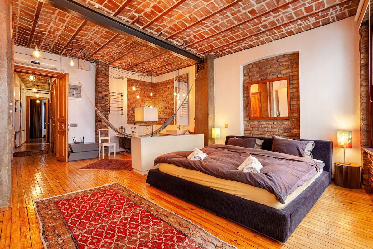 A bedroom with a large bed, wooden flooring, a red patterned rug, a brick wall, and a staircase.