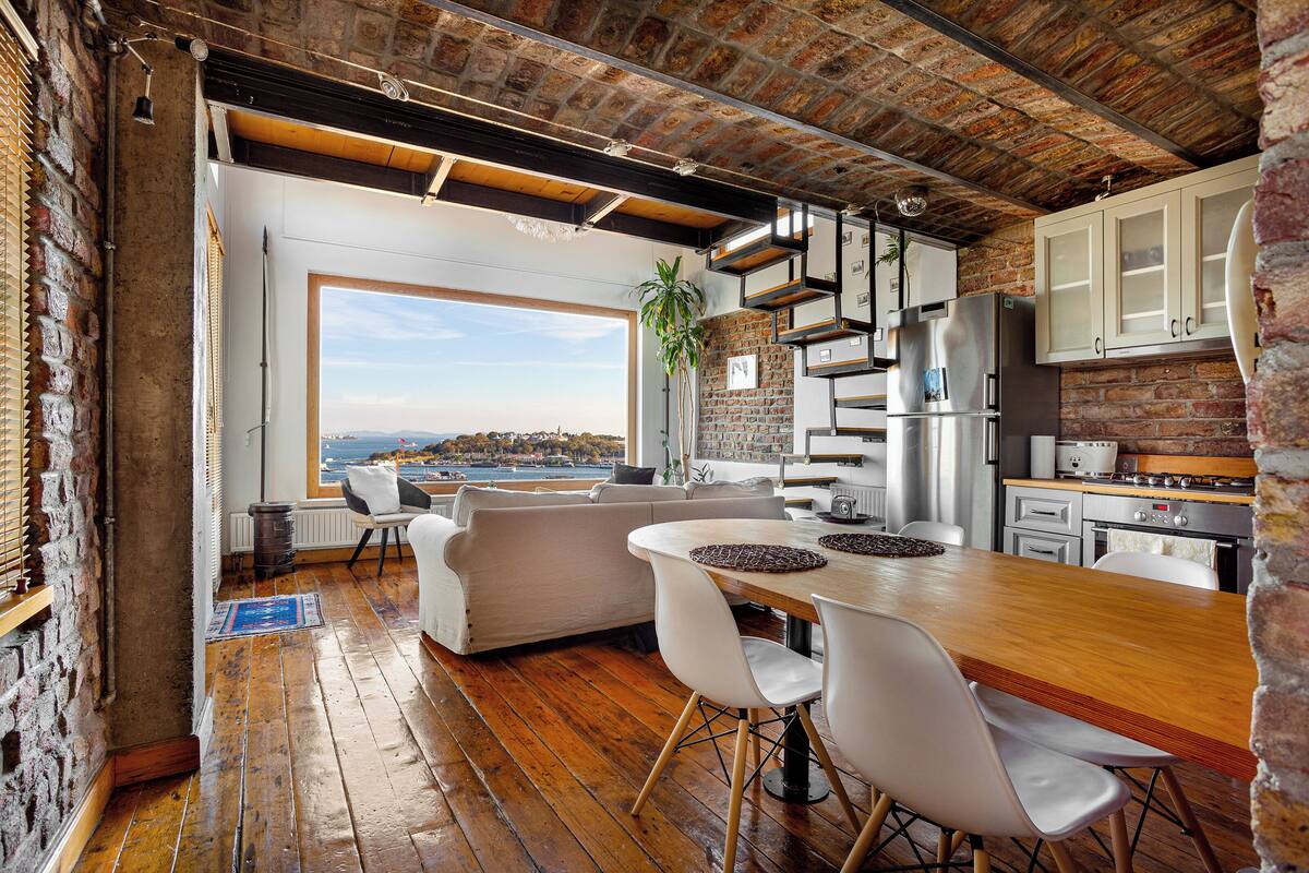 A modern kitchen with a wooden table and white chairs, a large window overlooking a scenic view, and a brick wall.