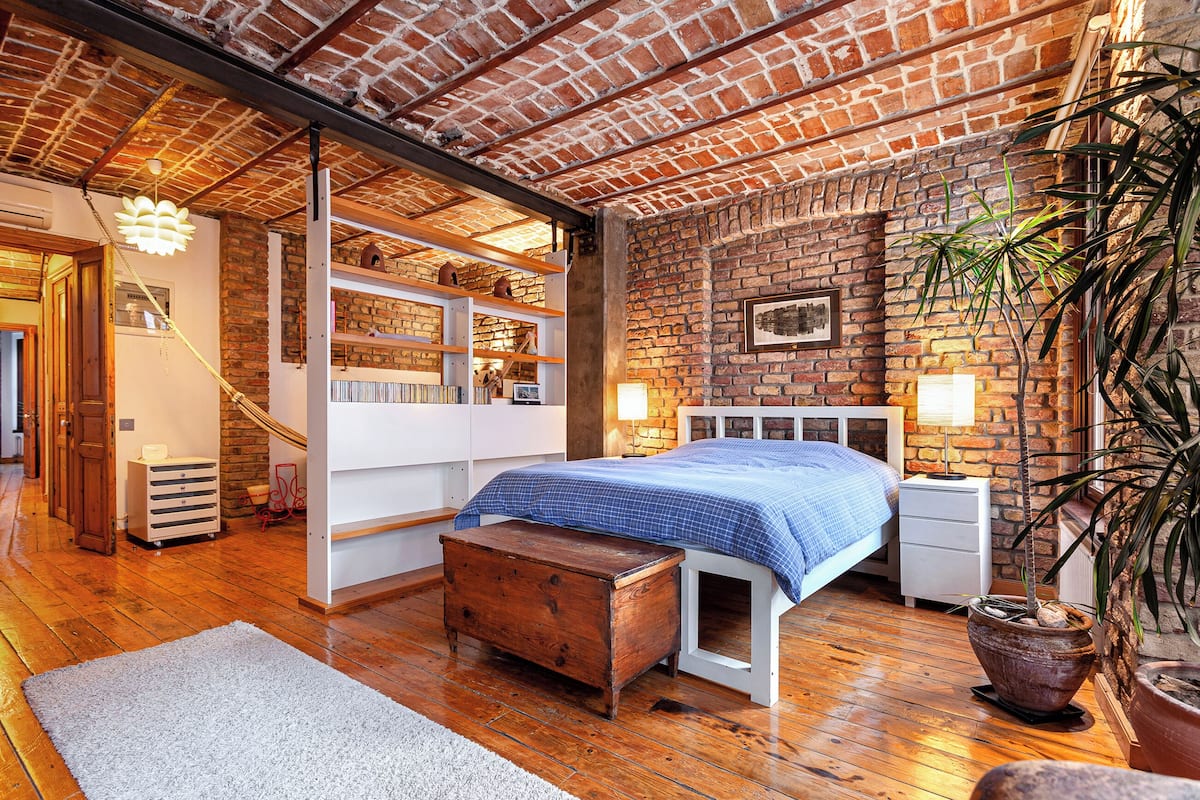 A bedroom with a brick wall, wooden floor, a bed with a blue bedspread, a wooden bench, a white bedside table, and a potted plant.