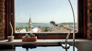 Blick aus einem Fenster mit Schreibtischlampe, zwei Topfpflanzen und freier Sicht auf eine Stadtlandschaft mit einem Turm und Wasser.